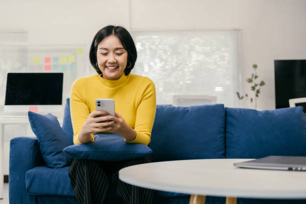 istock Young asian woman smiling while using her mobile phone, sitting comfortably on a blue sofa in a modern living room with a laptop on a nearby table 2194074887