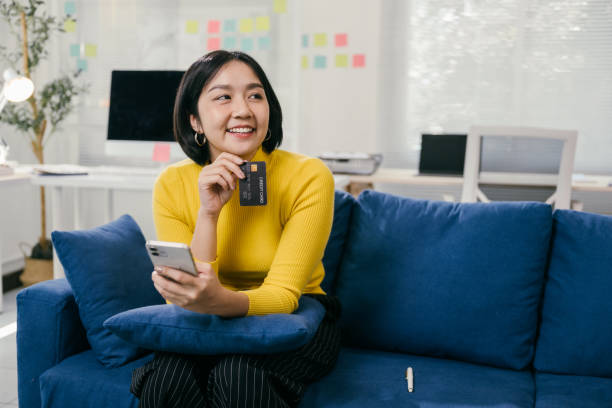 istock Happy young asian woman holding credit card and using smartphone while sitting on comfortable blue sofa in her living room, making online payment, shopping or banking 2194074908