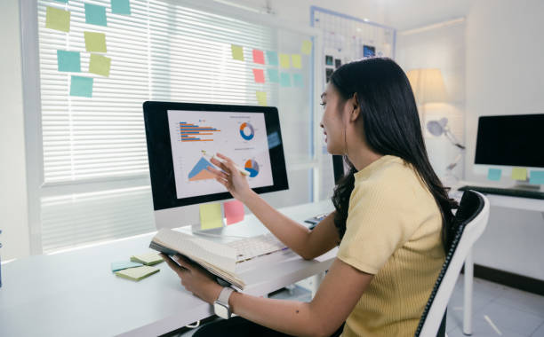 istock Young asian businesswoman pointing at financial charts displayed on computer screen while holding open notebook in modern office with sticky notes on window and wall 2194075279