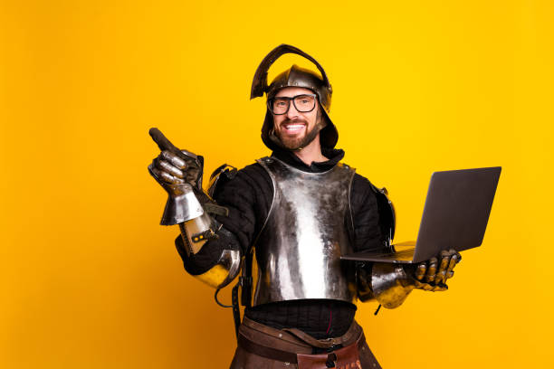 istock Young man wearing medieval knight armor holding a laptop with a cheerful expression against a bright yellow background 2194075795