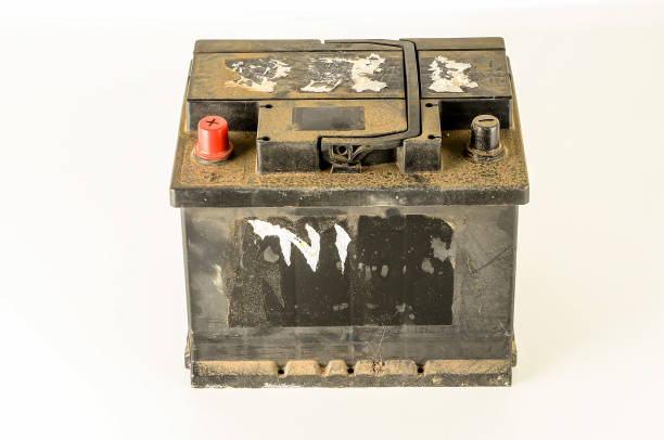 A dirty car battery sits on a white background. The battery is old and has a black cover