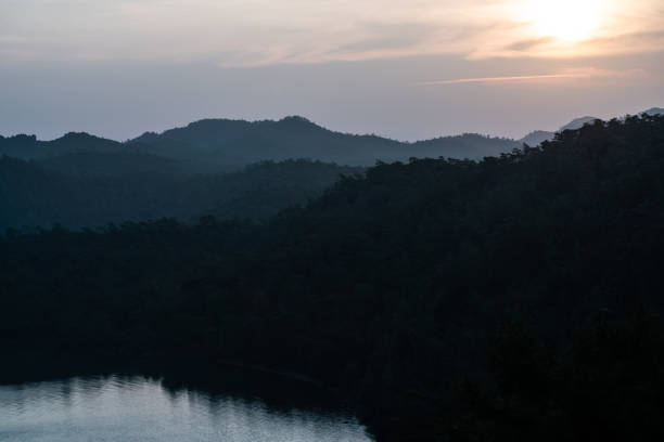 A tranquil evening scene capturing tree-covered hills with a peaceful reflecting lake below. The golden hues of the sky provide an ambient backdrop enhancing the serenity of the picturesque landscape.