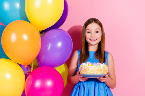 A cheerful young girl holding a decorated cake with colorful balloons against a pink background. The vibrant scene conveys joy, celebration, and a festive atmosphere perfect for young birthday events.