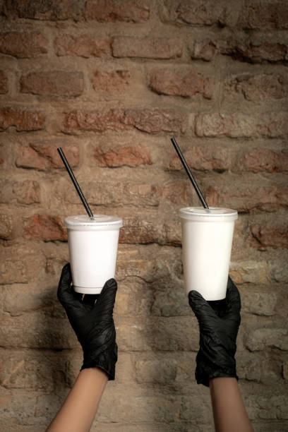 Two refreshing milkshake drinks served in clear plastic cups with black straws, Held by gloved hands, Against a rustic brick wall background
