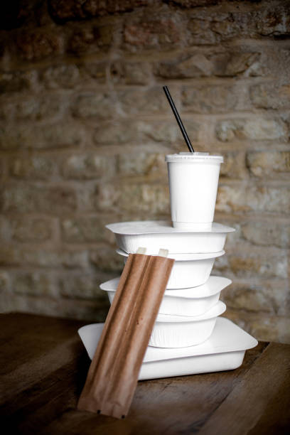 A neatly stacked collection of white takeaway food containers accompanied by wooden chopsticks and a refreshing drink in a cup with a striped straw, Placed against a rustic brick wall background