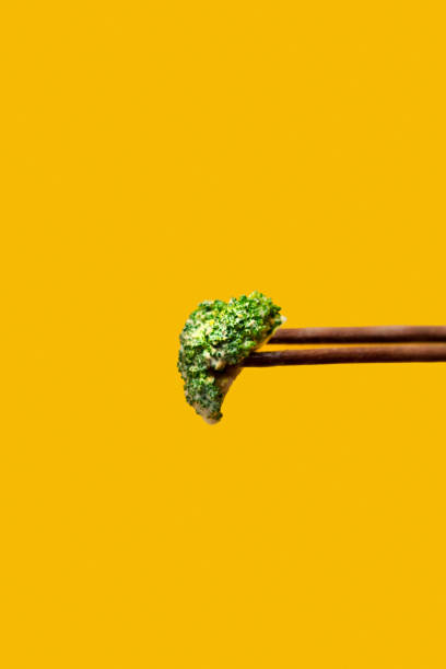 This striking image features a fresh green broccoli floret delicately held by wooden chopsticks, set against a bold yellow backdrop. The minimalist composition highlights the vibrant color contrast, making it ideal for campaigns promoting healthy eating, vegan lifestyles, or modern food aesthetics. Perfect for advertisements, recipe books, or educational materials on nutrition and sustainability.