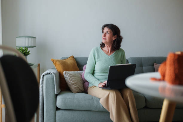 istock Senior adult woman using laptop computer sitting on sofa at home looking away 2194114596