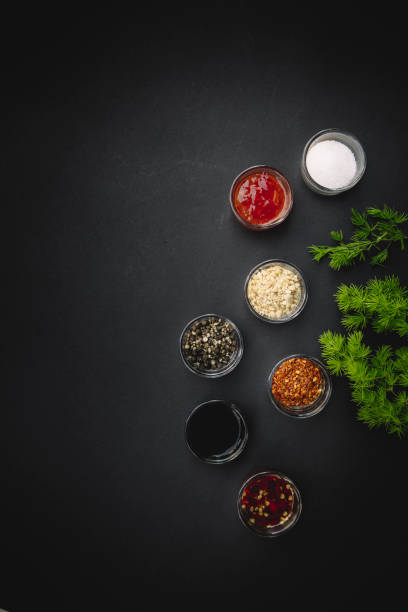 This image showcases a variety of condiments and spices beautifully arranged in small glass bowls on a dark background. The assortment includes soy sauce, chili flakes, sesame seeds, black pepper, and ketchup, accompanied by fresh parsley for a touch of greenery. The minimalist composition emphasizes the textures and vibrant colors of the ingredients, making it ideal for themes of cooking, seasoning, and food photography.