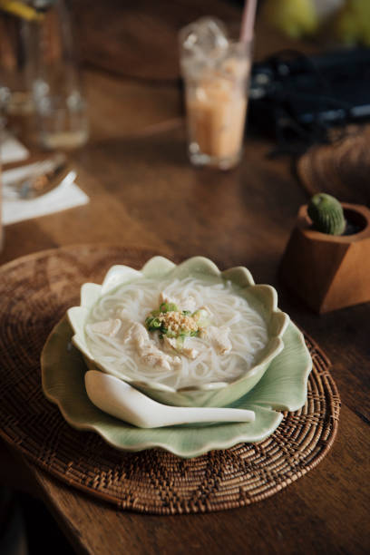 Traditional Thai Guay Teaw Gai noodle soup served in an elegant lotus-shaped bowl, Garnished with green onions and fried garlic, On a woven mat, Highlighting authentic Thai flavors
