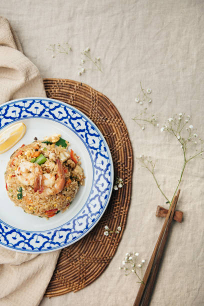 Traditional Thai fried rice with shrimp (Kung) and egg, Served on a blue and white patterned plate with a lemon wedge, Placed on a rustic woven mat and fabric backdrop, Showcasing authentic Thai cuisine This image highlights a plate of traditional Thai fried rice with shrimp (Kung) and egg, garnished with fresh vegetables and accompanied by a lemon wedge. The dish is beautifully presented on a decorative blue and white patterned plate, set atop a rustic woven mat and a beige fabric background. The natural and warm composition emphasizes the vibrant textures and colors of the fried rice, making it a perfect representation of authentic Thai flavors and homemade culinary art. jasmine rice stock pictures, royalty-free photos & images