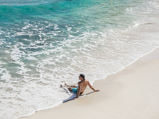 Shirtless man in swim trunks standing on sandy beach looking at calm ocean waves. Relaxed adult enjoying seaside view on sunny day. Beach vacation and summer leisure concept.