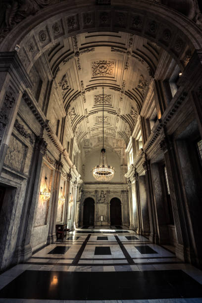 Amsterdam, Netherlands - July 2, 2018: This image captures the grandeur of the Amsterdam Royal Palace (Koninklijk Paleis) with its opulent neoclassical interior. The symmetrical hallway is adorned with intricately carved ceilings, elegant chandeliers, and marble walls. The soft lighting emphasizes the detailed ornamentation.