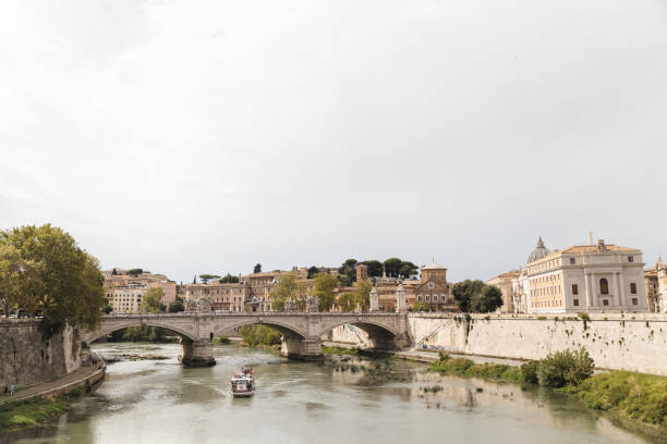 A tranquil scene featuring the Tiber River winding through the heart of Rome. The elegant Ponte Sant’Angelo bridges the river, framed by historic buildings and lush greenery. A boat glides peacefully along the water, adding a touch of life to this serene cityscape. The dome of St. Peter’s Basilica is visible in the distance, completing the picturesque Roman skyline.