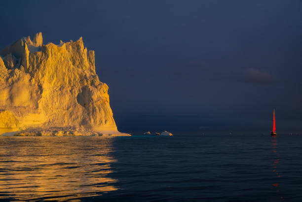 red sailboat sailing among floating yellow icebergs hit by setting sun in Disko Bay during polar summer midnight solar season. Ilulissat, Greenland