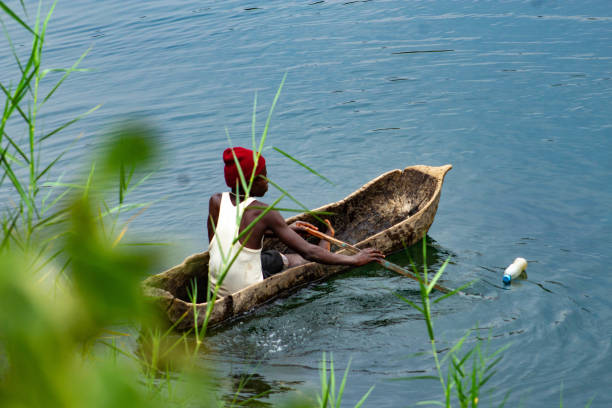 A fisherman maneuvers his traditional wooden canoe on the calm waters of Lake Kivu, Rwanda. The scene captures the simplicity of daily life and the enduring fishing practices in the region. Surrounded by lush vegetation, the image highlights the natural beauty and cultural significance of this rural setting. Editorial use only.