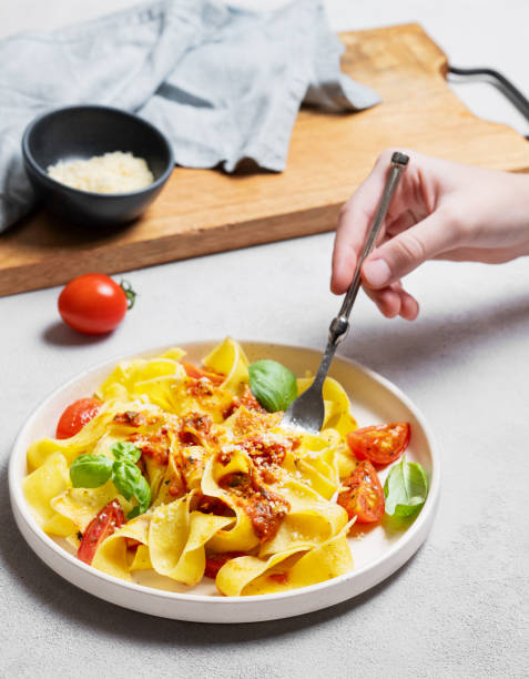 istock Hand holding fork with tagliatelle or pappardelle pasta with tomato sauce, basil and parmesan cheese in plate 2194174063