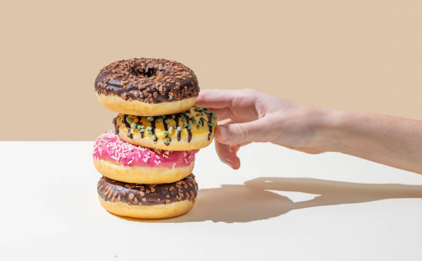 istock Hand takes one donut from stack of glazed donuts with different toppings on beige and white minimalistic background. 2194174253