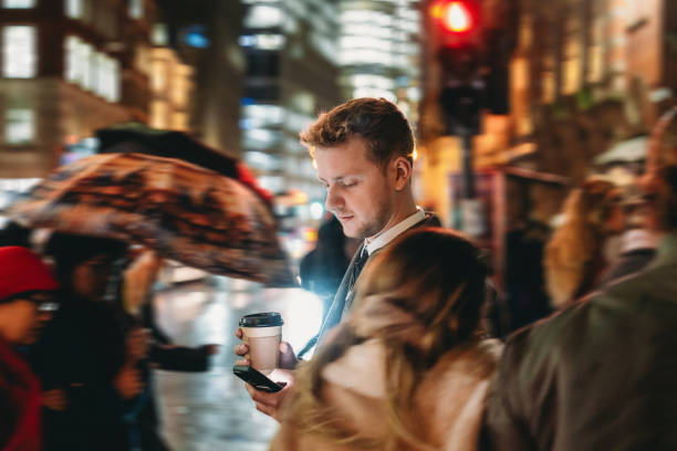 Young businessman is walking in the city at night, using his smartphone and drinking coffee, while people with umbrellas pass by quickly