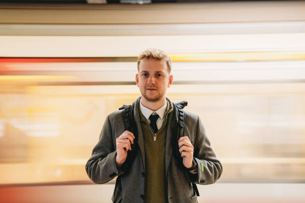 Young businessman with a backpack waiting on a platform for the train, surrounded by the bustling energy of the urban transportation scene