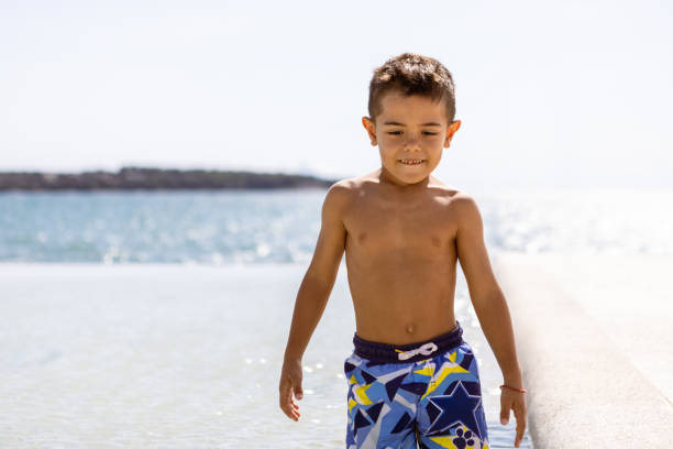 A young boy enjoying a sunny day at the beach, standing in front of the sea and wearing colorful swim trunks.