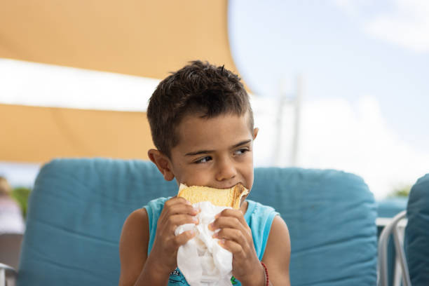 A young boy sits on a blue chair, happily biting into a sandwich while enjoying the outdoor ambiance.