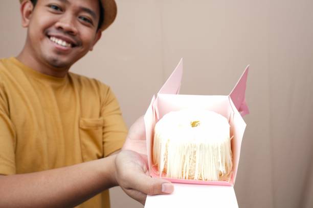 Smiling Asian man presenting a cheese-covered sponge cake with both hands.