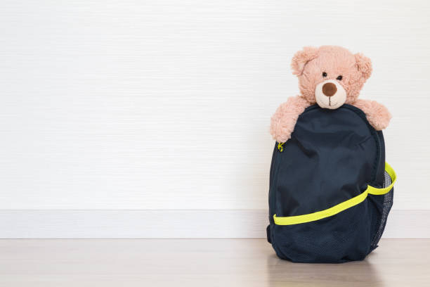 Smiling brown teddy bear looking behind opened dark school backpack on wooden floor at light gray wall background. Closeup. Empty place for text. Front view.