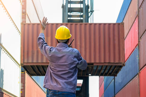 Dock supervisor in yellow hard hat and uniform directing container loading with walkie talkie and hand gesture at shipping container yard.