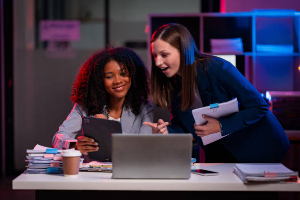 istock Two businesswomen working overtime, chatting in the office, working with laptops, documents, graphs, financial analysis with lights in the office 2194195394