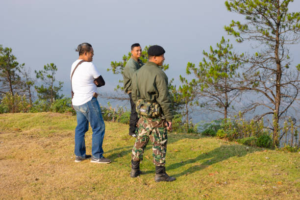 Three thai men looking in to mountain panorama landscape of Chiang Mai province from peak of Doi Mong Angkhet at morning. Men have different age. One is wearing jeans and has special thai hair cut. One of men is a soldier in uniform with camouflage pants, boots and a barrette. Men are talking and watching into landscape. In background are trees. Men are member of one of several groups of people visiting peak of mountain