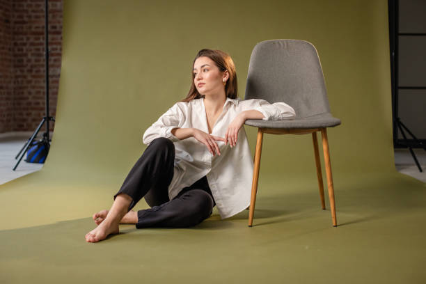 A modern woman poses thoughtfully with a chair in the studio. Dressed in a white shirt and black pants against a green background, she exudes calm and introspection.