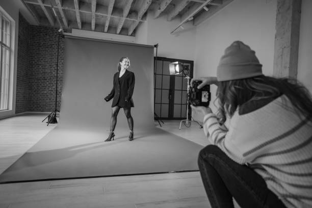 A photographer wearing a beanie captures a female model in a dynamic pose against a gray backdrop in a spacious studio. Natural light streams through large windows.