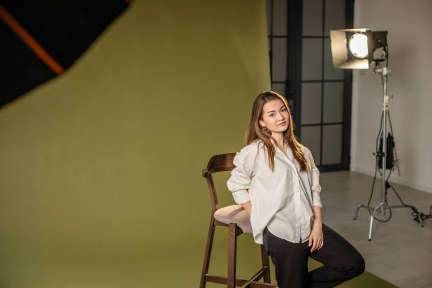 A woman in a white shirt and dark pants poses confidently against a green backdrop, with professional lighting equipment illuminating the studio setup.