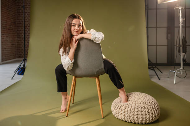 A woman sits barefoot on a chair placed backward, with a green backdrop in a professional studio, radiating a relaxed and contemplative mood.