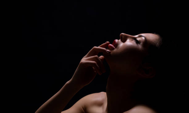 A woman poses gracefully in a dark environment, her face partially illuminated, conveying introspection. She gently touches her chin, lost in thought, showcasing elegance and emotion.