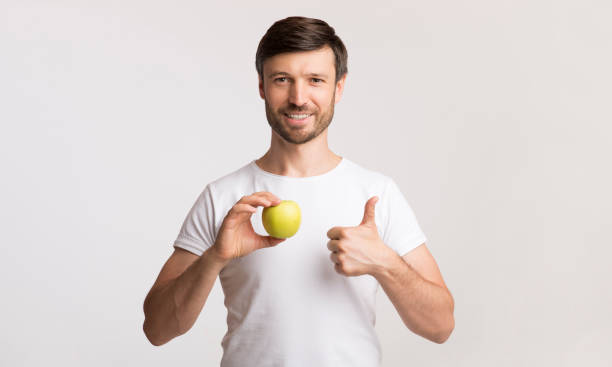 istock Man Smiles While Holding Green Apple and Giving a Thumbs up in a Neutral Background 2194236579
