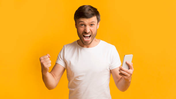 istock Excited Man Celebrating Success While Holding a Smartphone Against a Bright Yellow Background 2194237156
