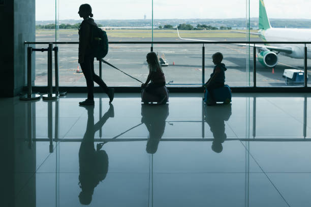 A mother pulls two children on small ride-on luggage through airport terminal, silhouetted against large windows with airplane in view. This scene captures family travel and fun moments before boarding a flight.