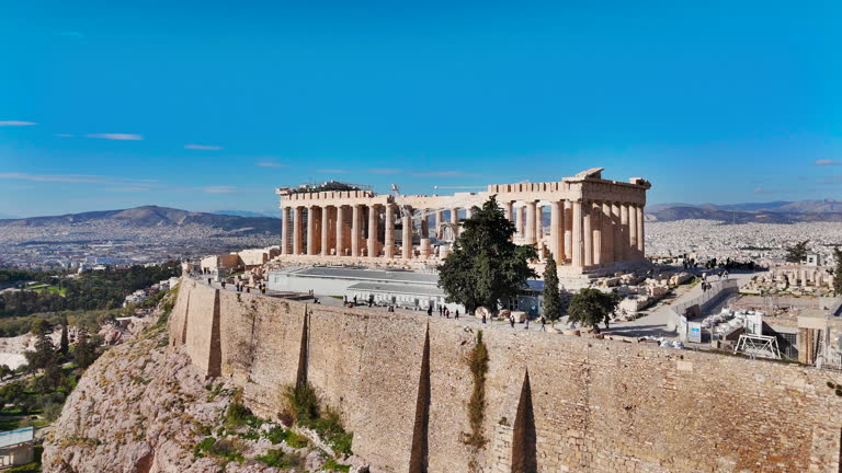 Aerial view of the Acropolis in Athens, Greece, Highlighting the massive stone fortifications, The Parthenon temple and the sprawling historic cityscape under a clear blue sky