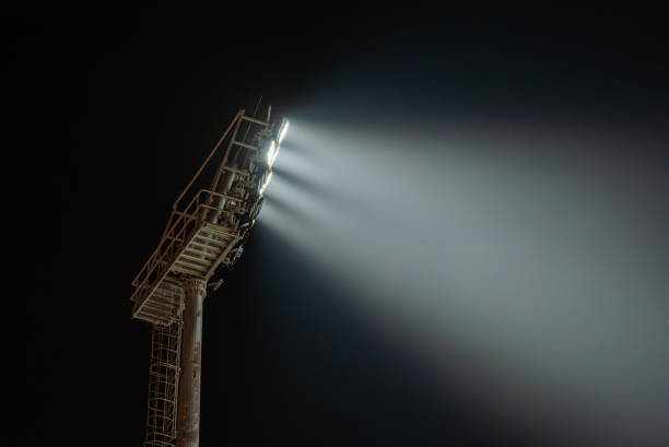 A tall light pole with floodlights shining beams of light against a dark night sky, surrounded by trees with autumn foliage