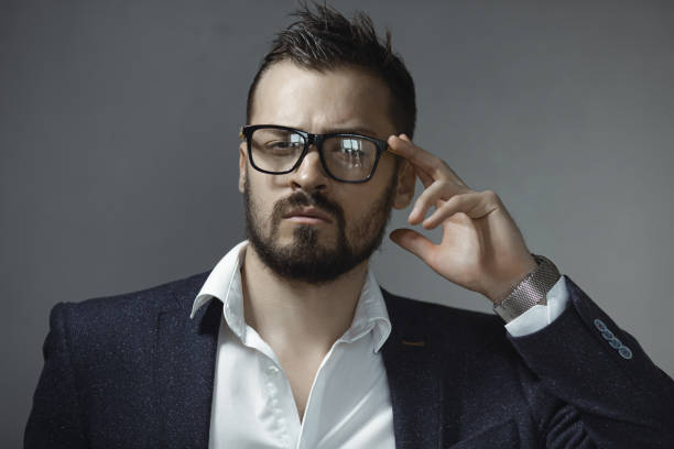 Serious young businessman wearing glasses and a formal suit, striking a thoughtful pose with a hand gesture. Perfect for leadership, corporate, and career-oriented photography themes.