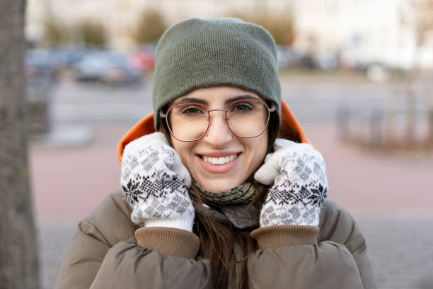Happy young woman wearing winter clothes, hat, gloves and eyeglasses, is smiling and protecting herself from cold in an urban park during a cold winter day
