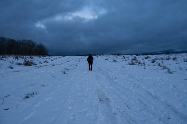 Tourist snow road. An unrecognizable man is walking along the road with a backpack. Dark blue saturated clouds. A winter atmospheric mood for walking in the countryside. Active lifestyle, walking