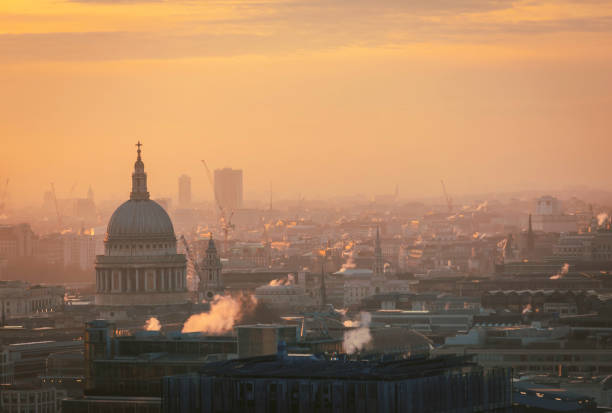 An atmospheric view of St. Paul’s Cathedral bathed in the golden light of sunrise, rising above the misty skyline of London. The historic dome stands tall amidst the modern cityscape, with construction cranes and office buildings framing the iconic landmark. Steam rises from rooftops, adding depth and a sense of early morning activity to the scene. The warm tones of the sky contrast beautifully with the cool architectural elements, capturing the essence of London’s blend of history and modernity