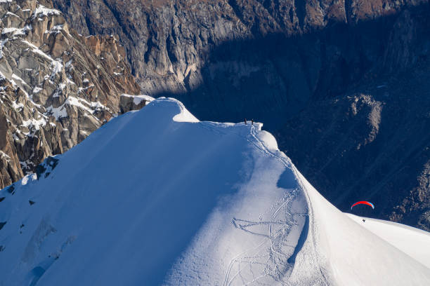 A snow-covered mountain peak with a paraglider soaring into a deep gorge leading to the Mer de Glace in Chamonix, as seen from Aiguille du Midi. A breathtaking alpine scene of adventure and freedom.