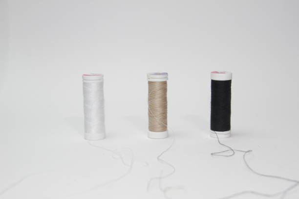 This image features three spools of thread arranged in a row on a white background. The spools are of different colors: white on the left, beige in the middle, and black on the right. Each spool has a loose end of thread extending from it, adding a dynamic element to the composition. This image is ideal for use in sewing, crafting, and textile-related projects, providing a clear and detailed view of the thread colors and textures.