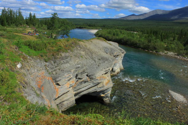 a gorgeous, mesmerizing view of the foothills of the Urals. a mountain river flows under a rock outcrop. bright day with white clouds on a blue sky. a group of tourists in the distance