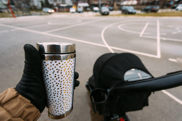 A hand wearing a glove holds a decorative thermos, overlooking a stroller in an urban park