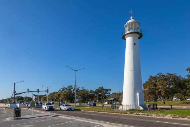 The historic Biloxi Lighthouse stands tall along Highway 90 on the Mississippi Gulf Coast. Built in 1848, this cast-iron lighthouse is a symbol of resilience, having survived many hurricanes, including Hurricane Katrina. The surrounding area features scenic coastal views, oak trees, and a busy roadway leading to Biloxi’s beachfront and casinos.