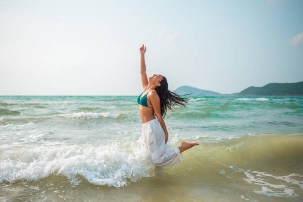 A young adult Hispanic female leaps with elation in shallow, clear waters on a sandy beach, dressed in swimwear under sunny skies.